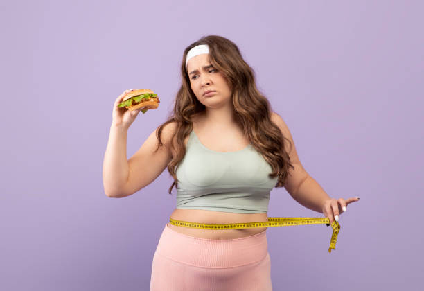 スポーツウェアの悲しいヨーロッパのミレニアル世代プラスサイズの女性は、測定テープでウエストを測定し、ハンバーガーを見ます - dieting sport exercising measuring ストックフォトと画像