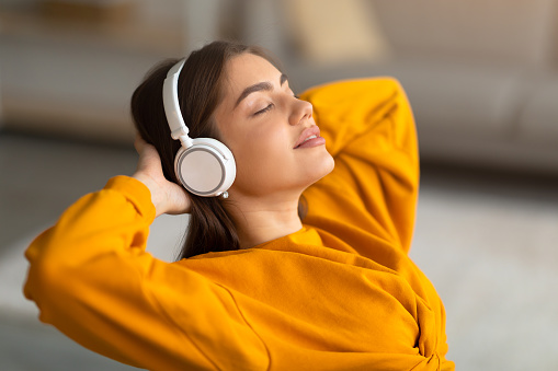 Calm young Caucasian woman listening to music in wireless headphones, relaxing with hands behind head at home. Lovely millennial female chilling with closed eyes and enjoying audio book