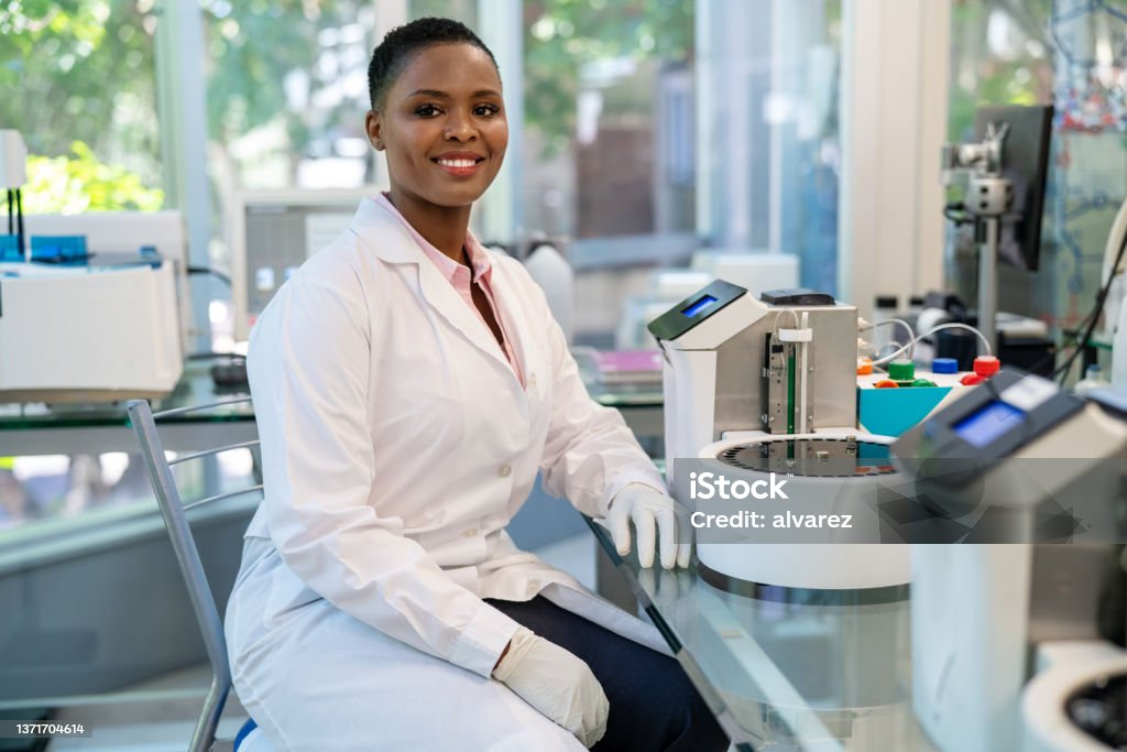 Portrait of African female scientist working in laboratory Portrait of African female scientist sitting on chair by lab equipment. Young doctor in lab coat working at laboratory. Medical Laboratory Stock Photo