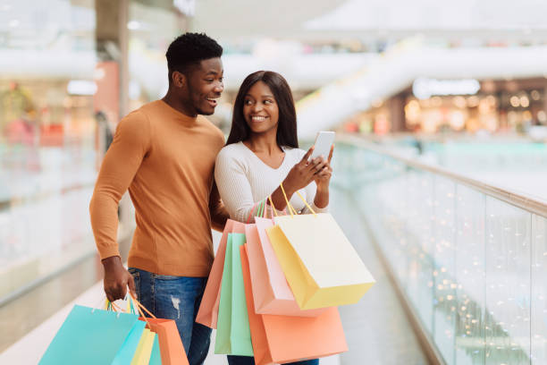portrait of excited black spouses using phone holding shopping bags - telephone indoors retail shopping mall imagens e fotografias de stock