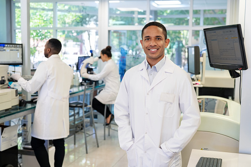 Portrait of a successful male scientist standing in lab with colleagues working in background. Medical researcher at science laboratory.