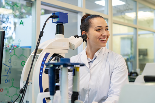 Happy female scientist sitting by microscope. Young woman doctor working at laboratory looking away and smiling.
