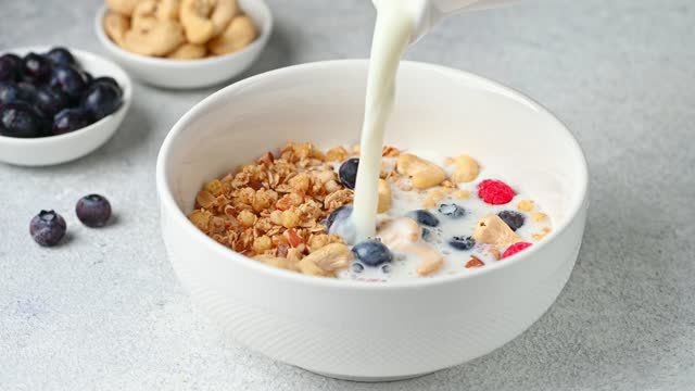 Pouring milk in white bowl with muesli cereals breakfast on white background. Granola with nuts and berries. Healthy breakfast. Making granola. Healthy food
