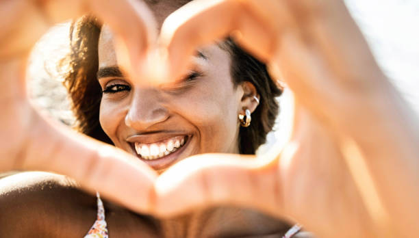 image en gros plan d’une femme souriante en maillot de bain sur la plage faisant une forme de cœur avec les mains - jolie femme hispanique joyeuse riant à la caméra à l’extérieur - mode de vie sain, amour de soi et concept de soins corporels - beautiful the human body body care close up photos et images de collection