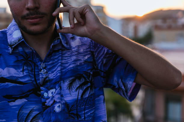 Unrecognizable young man in front with blue Hawaiian shirt talking on the phone with his smatphone during sunset. Concept: teleworking - cryptocurrency