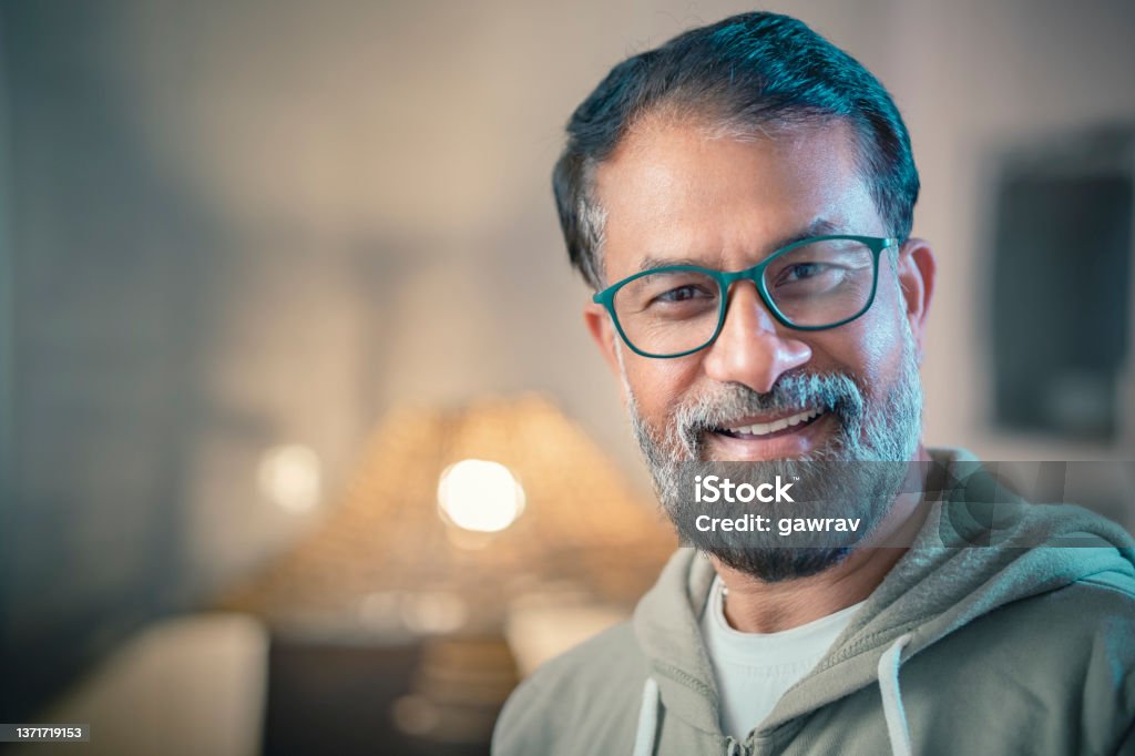 Mature man looks at the camera. In this headshot, indoor image, an Asian/Indian mature, happy man with a long beard looks at the camera with a toothy smile standing in his living room. He is in a warm hooded sweatshirt and eyeglasses. Portrait Stock Photo