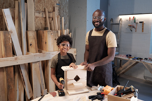 Portrait of happy black father building wooden birdhouse with son and looking at camera in workshop