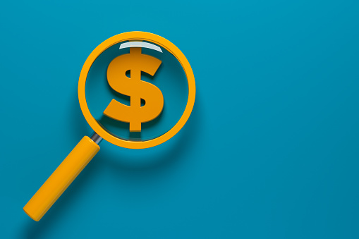 Orange-colored magnifying glass and orange-colored dollar symbol. On blue-colored background. Horizontal composition with copy space. No cutout.