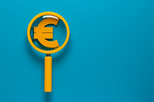 Orange-colored magnifying glass and orange-colored euro symbol. On blue-colored background. Horizontal composition with copy space. No cutout.