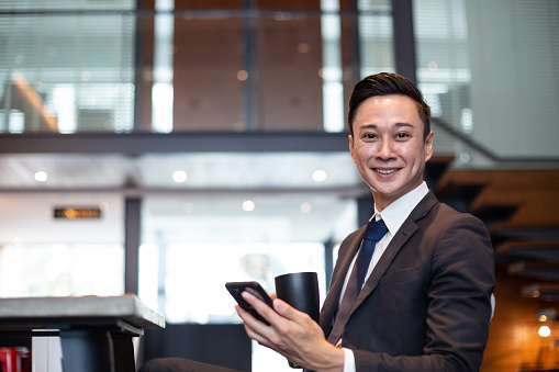 Young businessman holding a phone and smiling to camera while taking coffee break