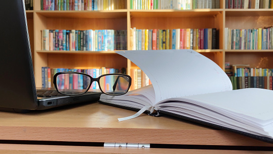Laptop, notepad and glasses on desk in library, bookshelf as background