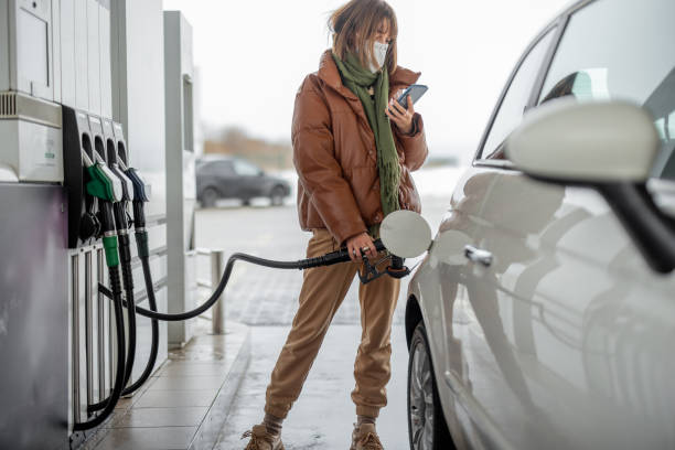 woman in face mask refueling car with a gasoline using smart phone - gas station fuel pump station gasoline imagens e fotografias de stock