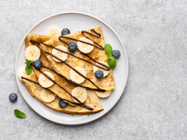 crêpes (dünne pfannkuchen, traditionelles russisches dessert blini) mit frischen blaubeeren, bananenscheiben, geschmolzener schokolade und grünen minzblättern auf keramikteller. - pancake blini russian cuisine french cuisine stock-fotos und bilder