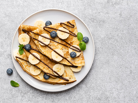 Crepes (thin pancakes, traditional russian dessert blini) with fresh blueberries, banana slices, melted chocolate and green mint leaves on ceramic plate.