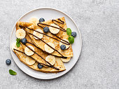 Crepes (thin pancakes, traditional russian dessert blini) with fresh blueberries, banana slices, melted chocolate and green mint leaves on ceramic plate.