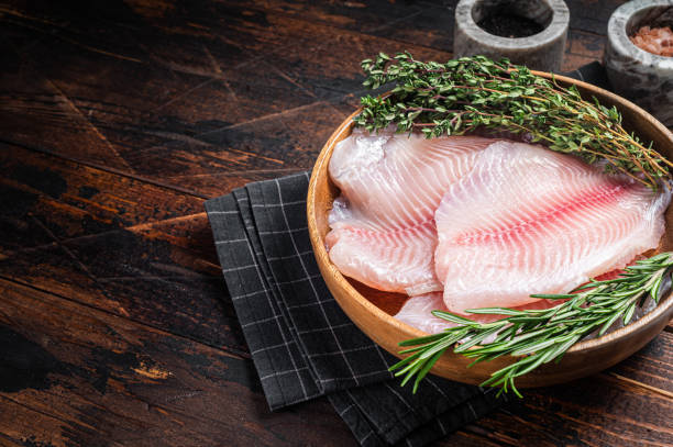 filetto di pesce bianco tilapia crudo in un piatto di legno con rosmarino e timo. sfondo in legno. vista dall'alto. spazio di copia - tilapia foto e immagini stock