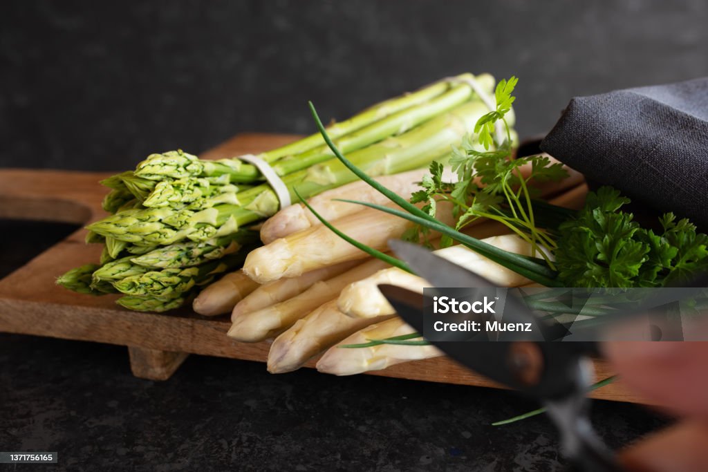 Fresh asparagus on wooden cutting board Fresh green and white asparagus on wooden cutting board against black stone wall. Vegetable for healthy nutrion concept. Lifestyle cooking background. Close-up with hort depth of field and space for text. Asparagus Stock Photo