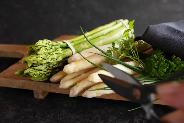 Fresh green and white asparagus on wooden cutting board against black stone wall. Vegetable for healthy nutrion concept. Lifestyle cooking background. Close-up with hort depth of field and space for text.
