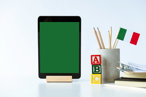 Front view of a white desk with a digital tablet, pen holder with pens and Italian flag, notepad,paperclip, and toy blocks with the letters A,B,C. Representing e-learning in Italy.
