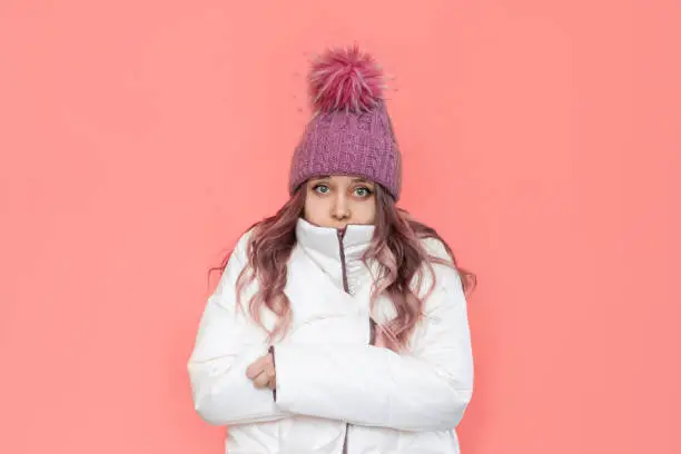 A young frozen caucasian pretty blonde woman in a white warm jacket and knitted hat shivers hugging herself isolated on a bright color pink background. Stylish winter clothes