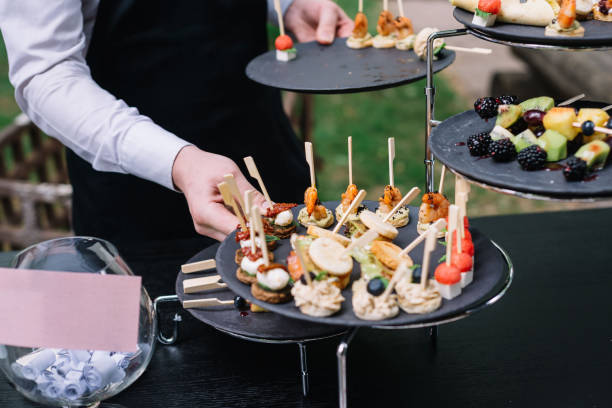 Waiter places plates on the table. Catering service. Wedding welcome food. Fruits on skewers and canapes. Welcome buffet at wedding reception. stock photo