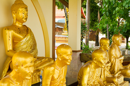 Golden buddha and thai monk statues at local temple in Sukothai province. Row of monks are sitting in front of buddha statue. In background are more temple buildings