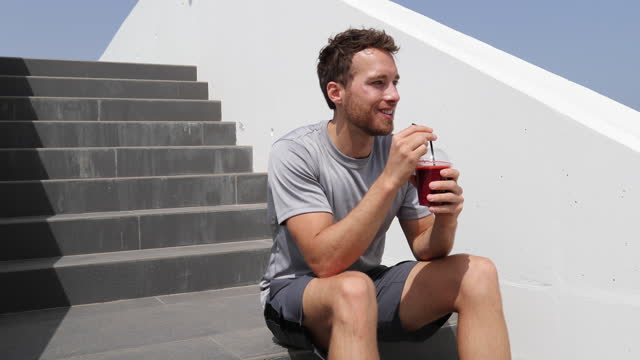 Juice smoothie sport man relaxing enjoying post workout morning breakfast sitting on outdoor grass at home or fitness gym. Athlete drinking red fruit smoothie drink.