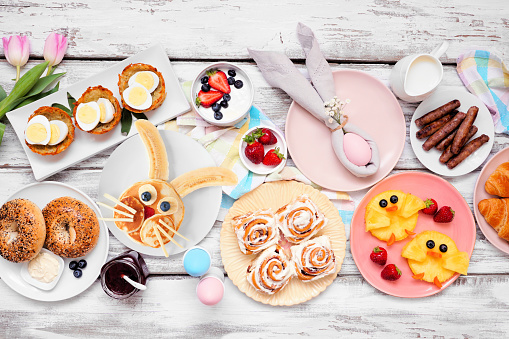 Easter breakfast or brunch table scene. Above view on a white wood background. Bunny pancake, egg nests, chick fruit and a variety of spring food items. Copy space.