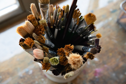 Above top close up view focus on paintbrushes in organizer standing on old retro painted table in artistic studio, working colored tools or used equipment of professional artist for artwork on canvas.