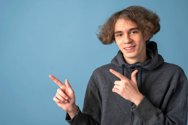 a smiling teenager in a gray hoodie and wavy hair points up with his hands on a blue background stock photo
