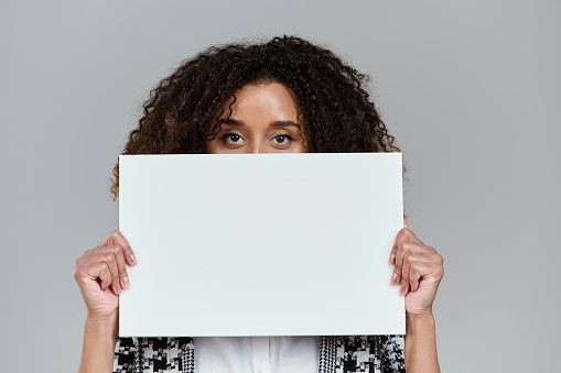 White cardboard to fill in messages. Blank copy space on sign. Office woman in studio shot. Light gray background. Mixed race model with Caucasian and American African background. Model showing excitement and happiness.