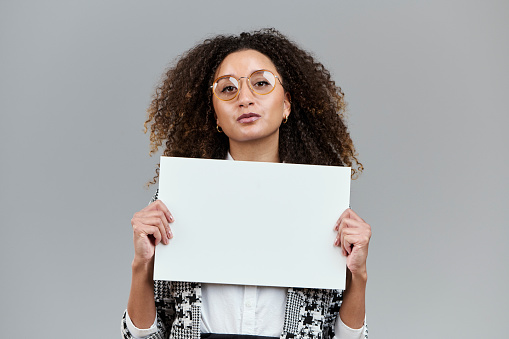 White cardboard to fill in messages. Blank copy space on sign. Office woman in studio shot. Light gray background. Mixed race model with Caucasian and American African background. Model showing excitement and happiness.