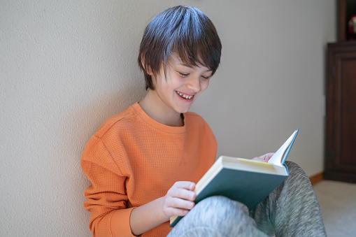 A happy tween boy sits on the floor in the living room of his home and reads a book. The adolescent is enjoying a quiet and relaxed morning, completely focused on the entertaining story.
