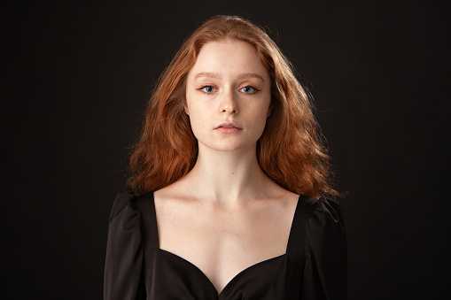 Close-up studio portrait of an attractive 20 year old red-haired woman in a black dress on a black background