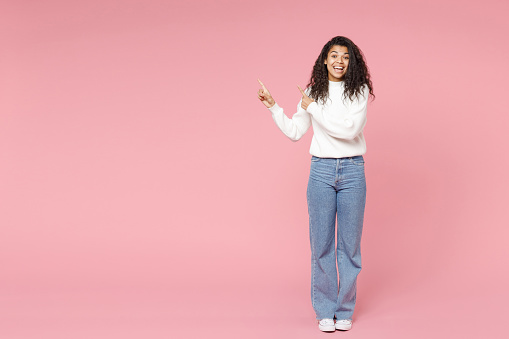 Full length young happy smiling african american curly woman 20s in white knitted sweater jeans point index fingers aside on copy space mock up area isolated on pastel pink background studio portrait