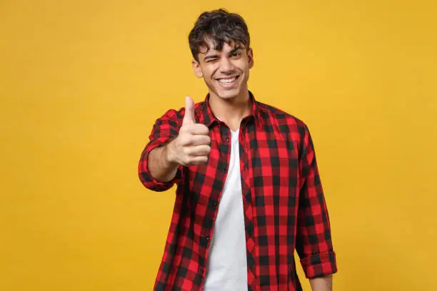 Young spanish latinos attractive handsome smiling stylish happy fashionable student man 20s wear red checkered shirt look camera show thumbs up gesture isolated on yellow background studio portrait