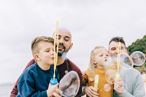 Gay male fathers and kids having fun outdoor playing with soap bubble toys - Lgbt and love family concept - Focus on left dad face
