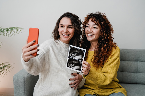 LGBT lesbian couple holding ultrasound photo scan on video call of growing baby in pregnancy time - Main focus on right woman face