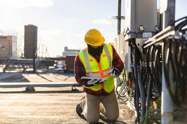 A blue-collar worker, a repairman, is checking and servicing telecommunication equipment mounted on a rooftop.