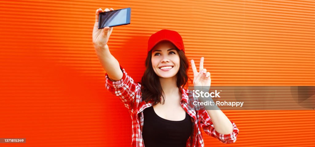Summer portrait of happy smiling young woman taking selfie by smartphone wearing baseball cap on red background Cap - Hat Stock Photo