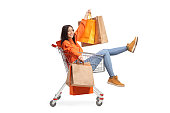 Full length portrait of a happy young female sitting inside a shopping cart and holding shopping bags