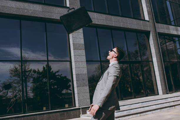 Side view young overjoyed smiling employee business man 20s in grey suit stand near office glass wall building outdoors in downtown city hold briefcase toss up diplomat bag look aside spread hands. vista lateral joven encantado empleado sonriente hombre de negocios de 20 años con traje gris parado cerca de la pared de vidrio de la oficina edificio al aire libre en el centro de la ciudad sostener maletín tirar bolsa de diplomático mirar a un lado m