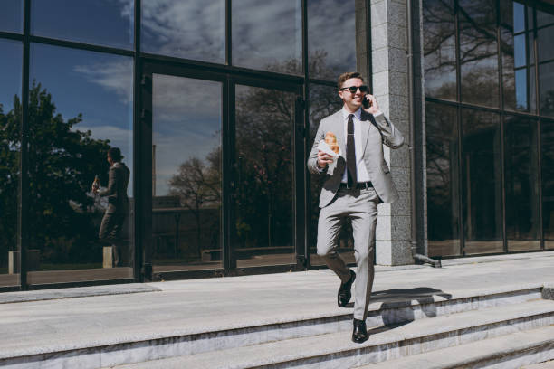 un jeune employé caucasien à succès homme d’affaires de 20 ans en costume gris monter sur les marches d’un bâtiment de mur de verre de bureau à l’extérieur dans le centre-ville du centre-ville en mangeant un hamburger fast-food déjeuner pendan - suit full length businessman 20s photos et images de collection