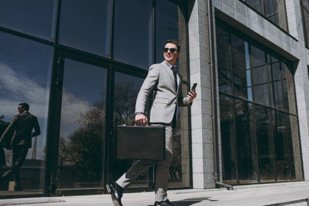 vue complète en bas jeune employé prospère homme d’affaires 20s porter costume gris marcher aller près du bureau mur de verre bâtiment à l’extérieur dans le centre-ville tenir téléphone portable porte-documents sac diplomate sac - suit full length businessman 20s photos et images de collection