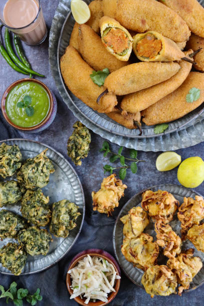 Stock photo showing a close-up, elevated view of a batch of homemade, fried mirch pakora (green chilli fritters) stuffed with an aloo mix, palak pakora (spinach fritters) and crispy, deep fried onion bahjees. imagen de la bandeja con plato estampado de buñuelos de chile verde caseros (mirch pakora), plato crujiente de cebolla bhajis y palak pakora (buñuelos de espinacas), vaso chai bowl menta y cilantro chutney, cocina india bocadillo vegetariano, fondo gris,