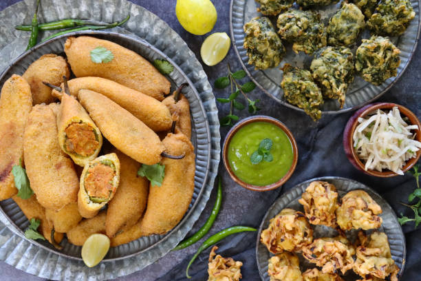 Stock photo showing a close-up, elevated view of a batch of homemade, fried mirch pakora (green chilli fritters) stuffed with an aloo mix, palak pakora (spinach fritters) and crispy, deep fried onion bahjees. imagen de la bandeja con plato estampado de buñuelos de chile verde caseros (mirch pakora), plato crujiente de cebolla bhajis y palak pakora (buñuelos de espinacas), tazón de menta y chutney de cilantro, bocadillo vegetariano de cocina india, fondo gris