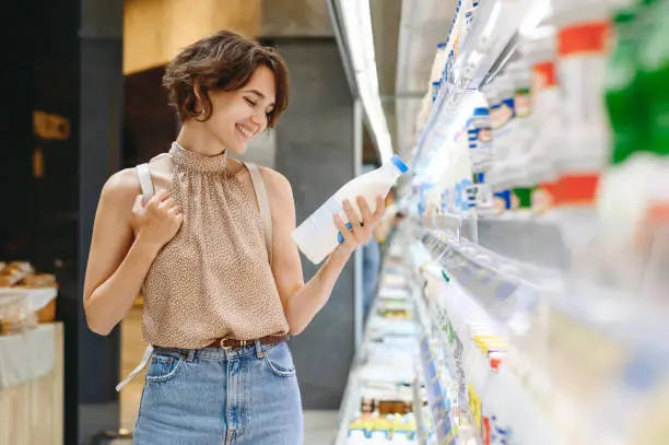 Photo of Young smiling happy woman 20s in casual clothes backpack shopping at supermaket grocery store buy choose dairy produce take milk read shelf life inside hypermarket Purchasing gastronomy food concept