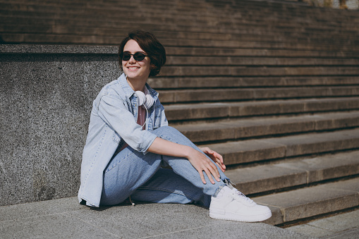 Side view full length young woman in jeans clothes headphones eyeglasses listen to music leaning on building wall sit on concrete steps outdoors walk look aside rest. People urban lifestyle concept