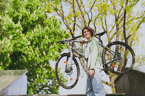 Young sporty strong fun happy woman 20s wear casual green jacket jeans holding bicycle bike strolling in city spring park outdoors, look camera. People active urban healthy lifestyle cycling concept.