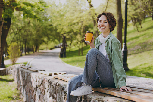 Young smiling fun woman in casual green jacket jeans sit on bench in city spring park outdoors hold takeaway delivery craft paper brown cup coffee to go, rest in morning People urban youthful concept